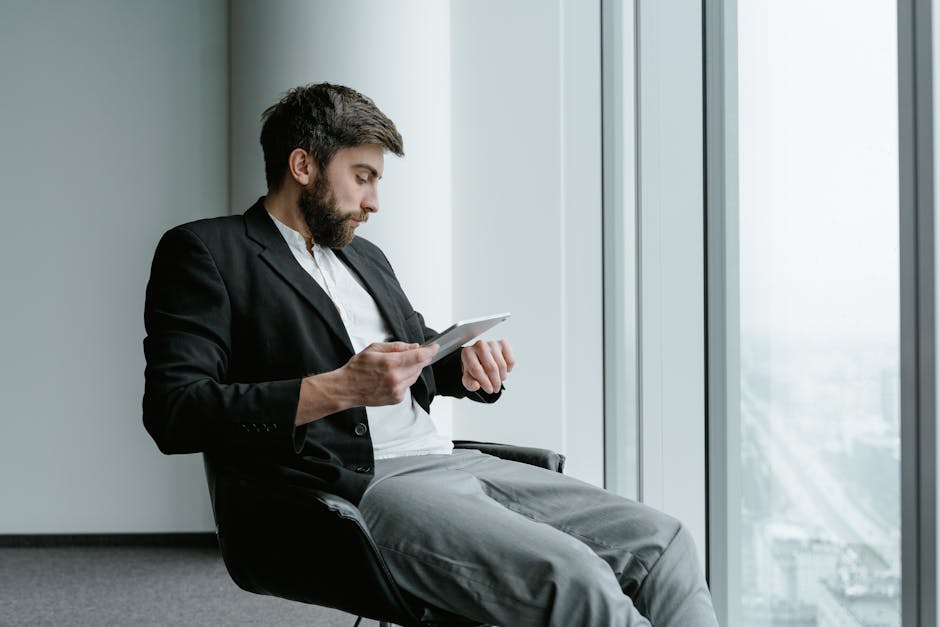 A businessman in a suit checks his watch while using a tablet in a modern office.